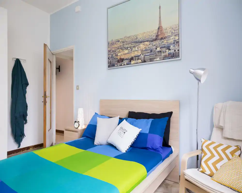 Bright, modern bedroom with a full double bed, colorful bedding, bedside table and wall art of the Eiffel Tower — clean, well-lit and tastefully styled for rental listings.