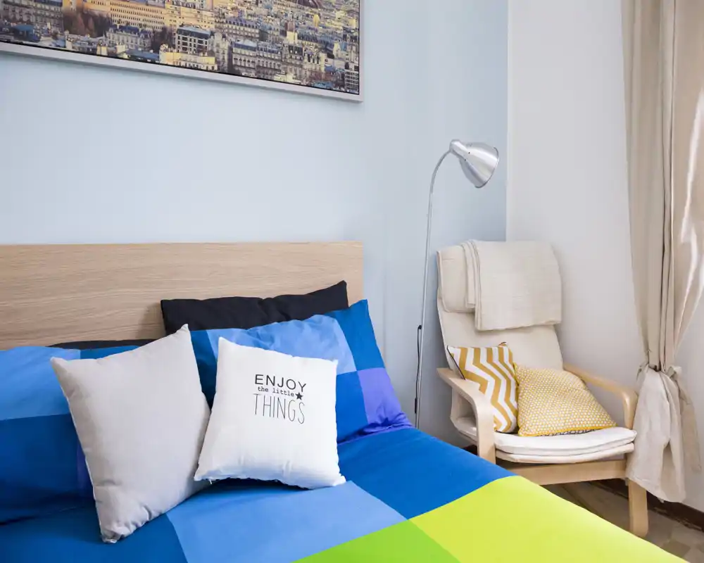 Bright, neatly styled bedroom with a colorful blue-and-green bedspread, decorative pillows and a cozy armchair by the window—ideal for rental photos.