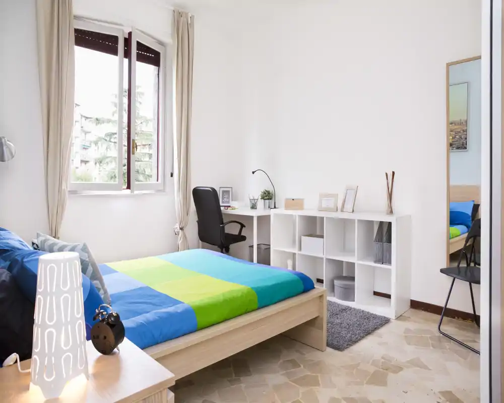 Sunny, neatly styled bedroom with a double bed, colorful duvet and a small desk by the window — bright and welcoming space ideal for relaxing or working.