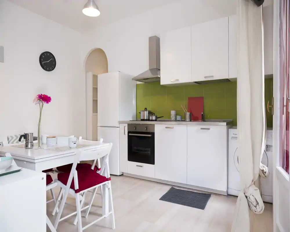 Compact modern kitchen with a white cabinetry, green tiled backsplash, built-in oven and full-height fridge. A small dining table with chairs and decorative touches (flower, cushions) creates a cozy, well-lit space ideal for short-term rentals.