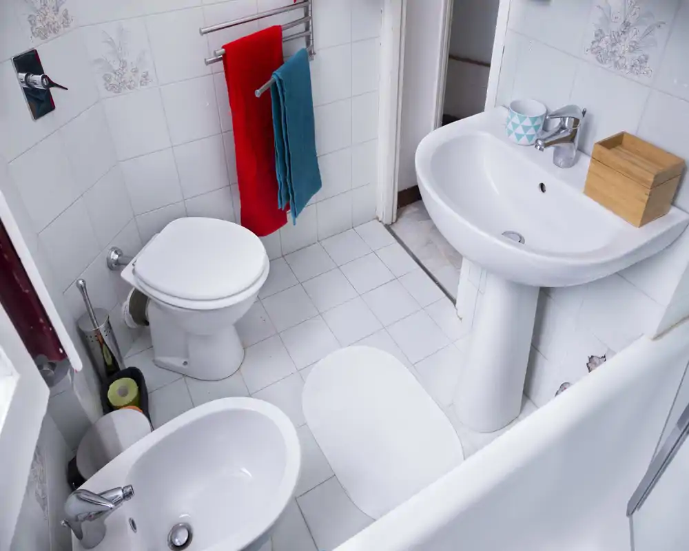 Clean, compact bathroom with white tiles, pedestal sink, toilet, bidet and a bathtub edge visible; bright and tidy space with colorful towels adding contrast.