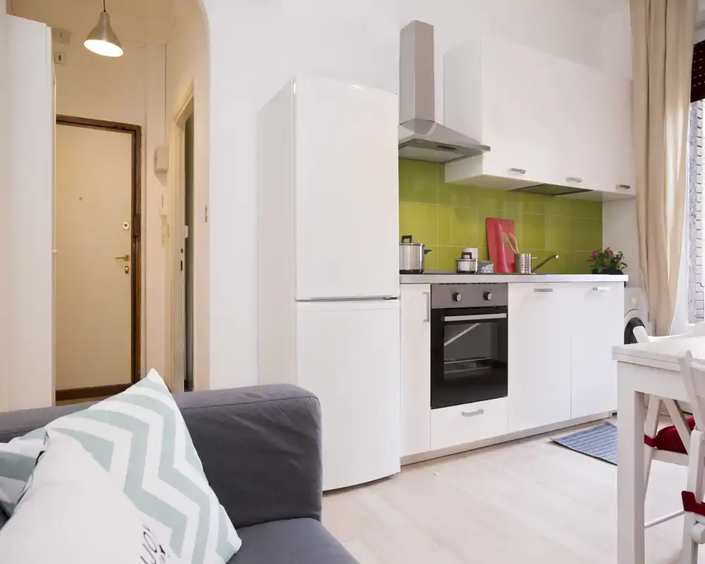 Bright, modern open-plan living area with a compact white kitchen, oven and fridge next to a cozy sofa — ideal for a small apartment or studio.