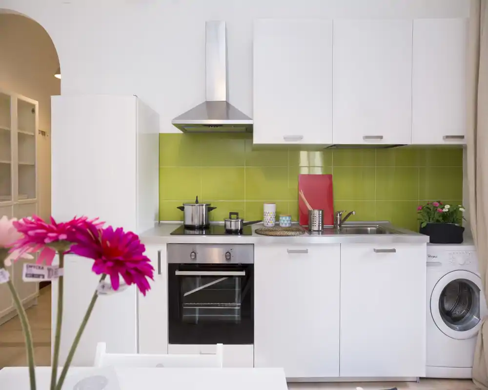 Modern compact kitchen with white cabinets, green tiled backsplash, oven and washing machine; bright, tidy and well-styled for a small apartment.