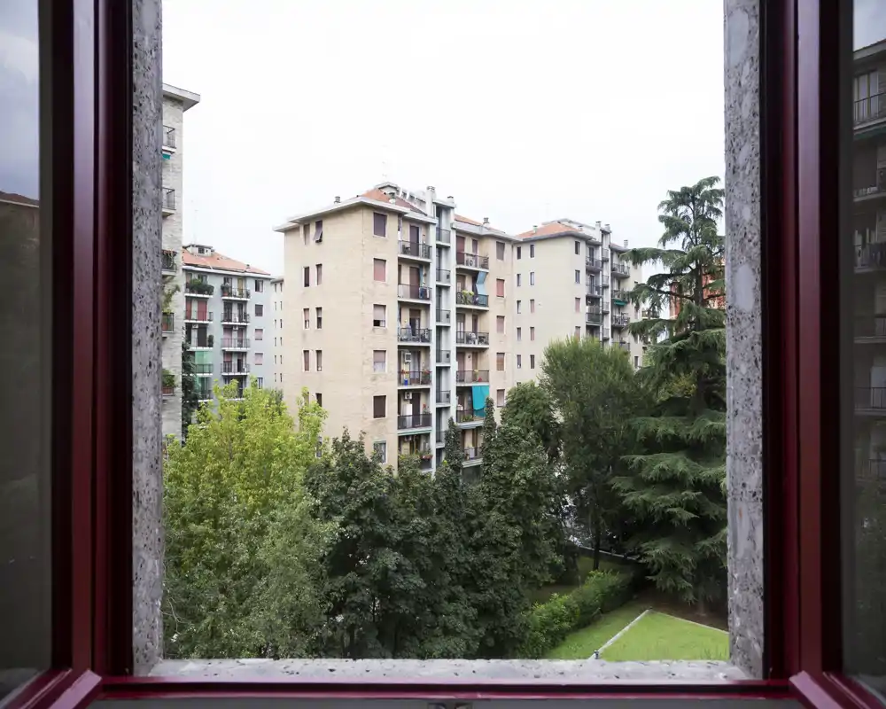Window view overlooking a residential courtyard with mid-rise apartment buildings and mature trees, offering a pleasant city-green outlook.