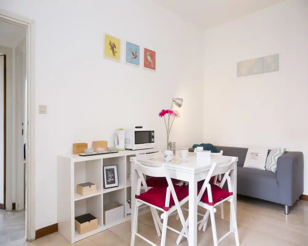 Bright, compact living/dining corner with a small sofa, dining table and shelving — styled with cushions, flowers and wall art for a cozy feel.