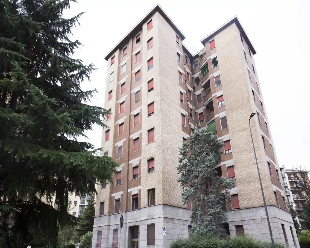 Exterior view of a multi-story brick apartment building with trees in the foreground and red window shutters, photographed from street level.