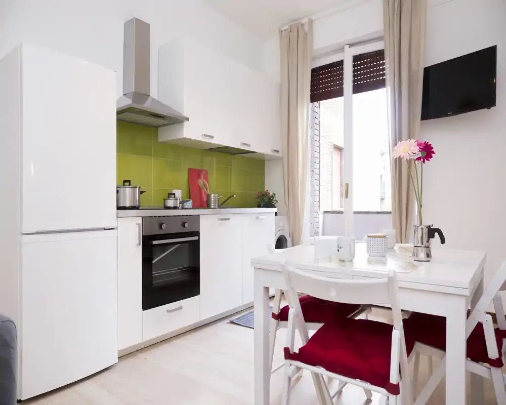 Bright modern kitchenette with white cabinets, integrated oven and a small dining table with red cushions — a compact, well-lit space ideal for short stays.