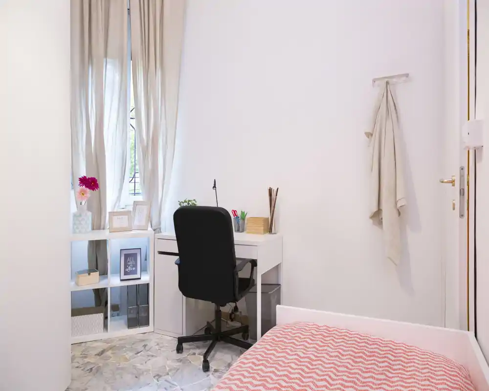 Small, bright bedroom with a single bed, a neat white desk and a compact shelving unit by the window — ideal for a student or remote worker.