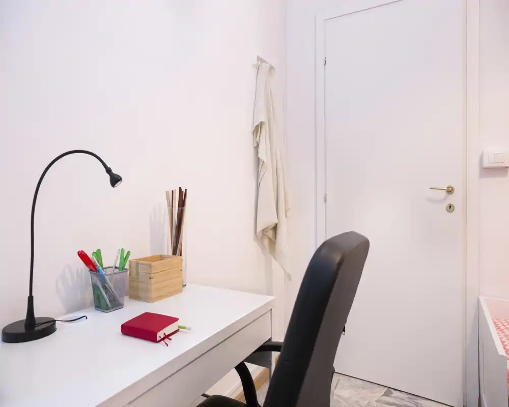 A neat study corner with a white desk, desk lamp and office chair. Simple, well-lit workspace with organized stationery and a soft towel hanging on the wall.