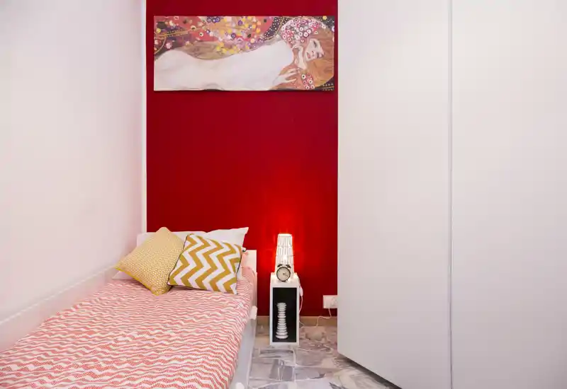 Bright, compact bedroom with a single bed against a deep red accent wall, patterned bedding and decorative cushions, plus a small nightstand with lamp and clock.