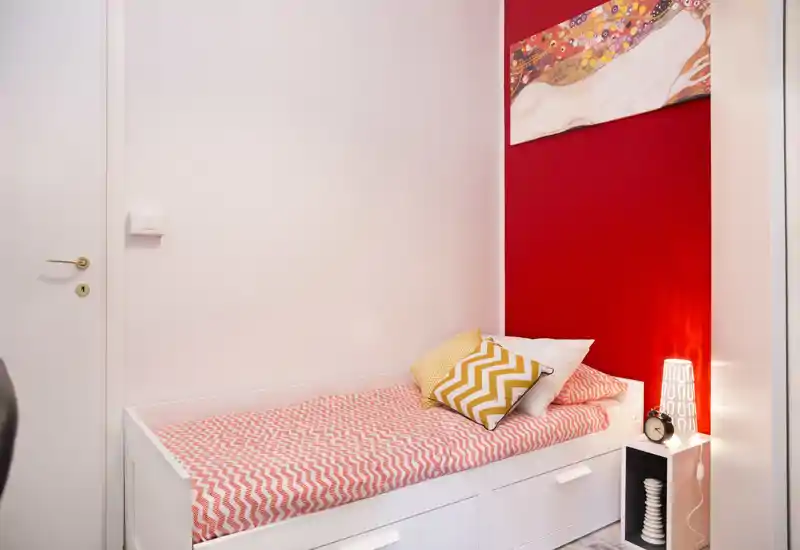 Small styled single bedroom with a daybed against a red accent wall, patterned bedding and decorative cushions; includes a bedside shelf with a lamp and alarm clock.
