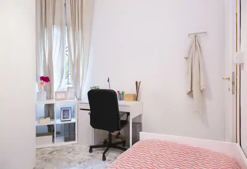 Small, bright bedroom with a single bed, a neat white desk and a compact shelving unit by the window — ideal for a student or remote worker.