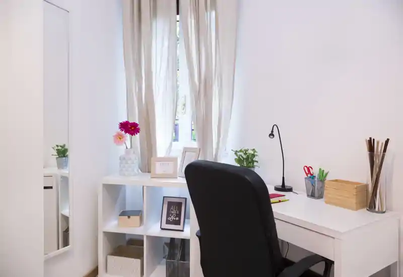 Bright, minimalist home office corner with a white desk, black ergonomic chair, shelving unit and decorative accents by a curtained window.