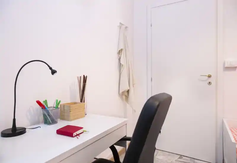 A neat study corner with a white desk, desk lamp and office chair. Simple, well-lit workspace with organized stationery and a soft towel hanging on the wall.