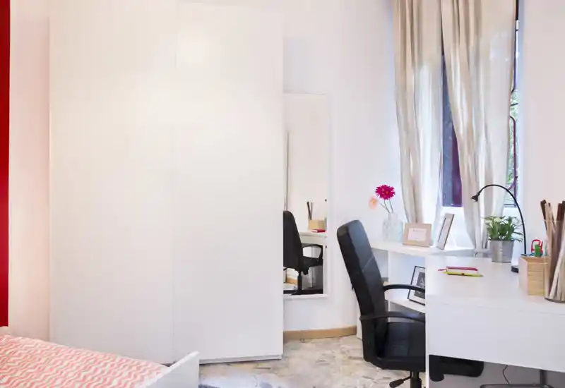 Bright, modern bedroom corner with a white wardrobe, a desk by the window and a partially visible bed with patterned bedding.