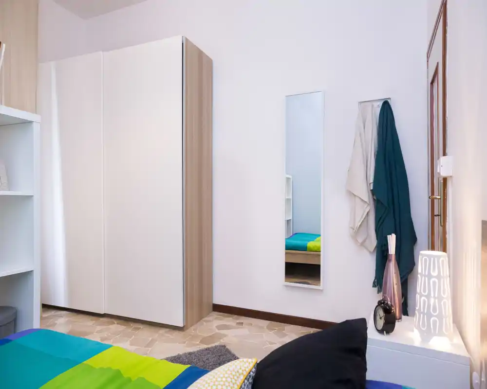 Dormitorio luminoso y ordenado con colcha colorida, armario y espejo de pie; lámpara de mesita y almacenamiento cuidado hacen la estancia acogedora.