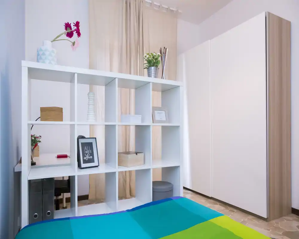 Dormitorio luminoso y bien decorado con una cama colorida en primer plano y una estantería modular que hace de separador; cortinas claras y un gran armario completan la estancia.