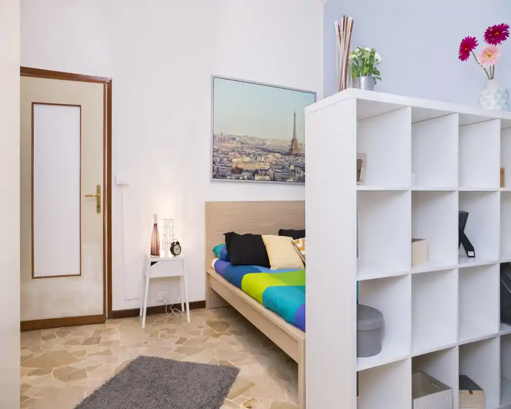 Dormitorio moderno y luminoso con cama colorida, mesita de noche y estantería decorativa que divide parcialmente el espacio; estilo limpio y acogedor para anuncios de alquiler.