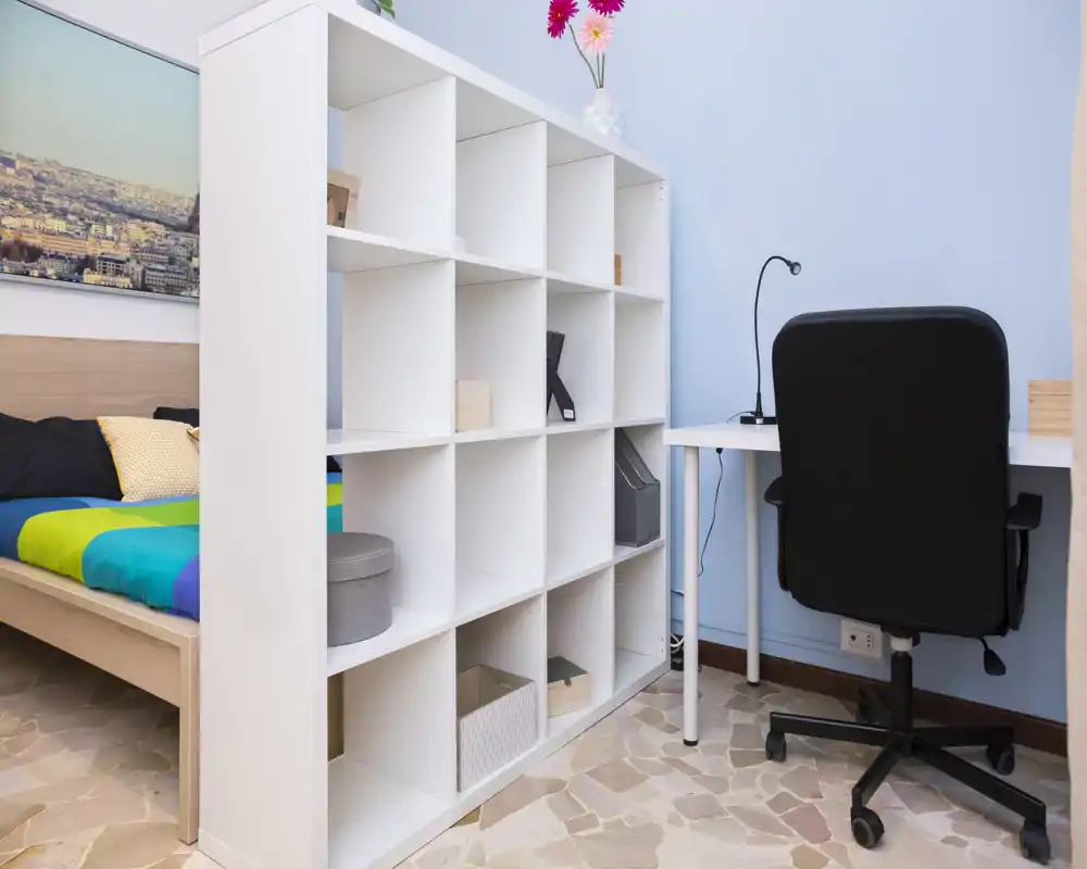 Habitación luminosa y ordenada con una cama colorida parcialmente visible detrás de una estantería blanca de cubos y un escritorio compacto con silla ergonómica — ideal para estudiante o una persona sola.