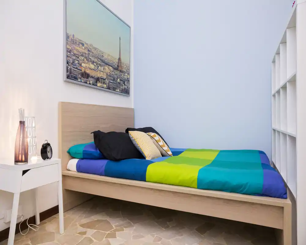 Dormitorio moderno y compacto con cama de madera, edredón colorido y cojines decorativos. Espacio luminoso y ordenado con mesita y estantería.