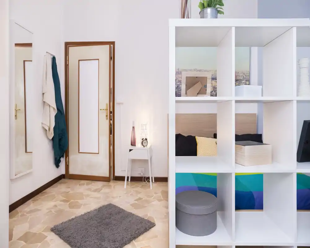 Dormitorio moderno y luminoso parcialmente visible detrás de una estantería blanca; textiles acogedores, cojines y una lámpara de mesita crean un ambiente atractivo para un anuncio de alquiler.
