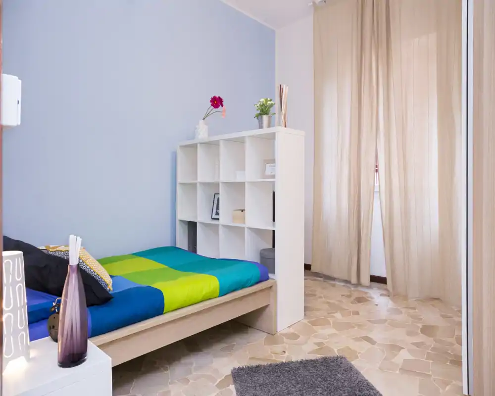 Habitación individual luminosa y bien decorada con cama colorida, estantería a modo de cabecero y luz natural suave desde la ventana con cortina.