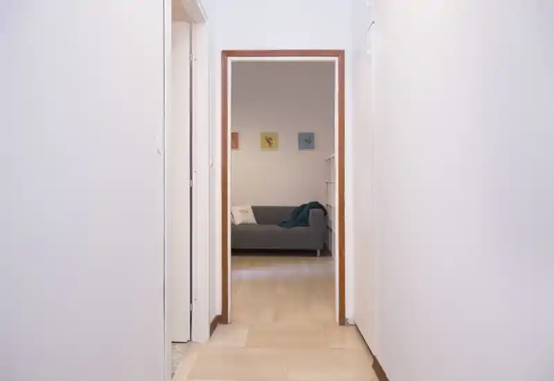 View down a clean hallway into a small living area with a grey sofa, throw and cushions; simple, neutral décor and soft natural light.