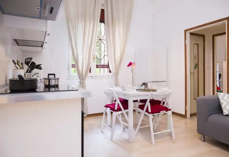 Bright, well-staged living area with a small dining table and compact kitchenette — airy curtains, decorative cushions and fresh flowers create a cozy, inviting atmosphere.