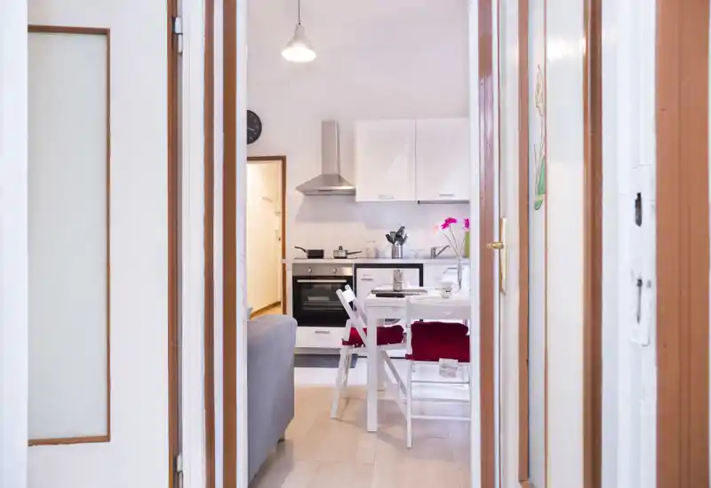 Bright, compact open-plan kitchen with white cabinets, a built-in oven, extractor hood and a small dining table set for two — tastefully staged with red chair cushions and a vase of flowers.