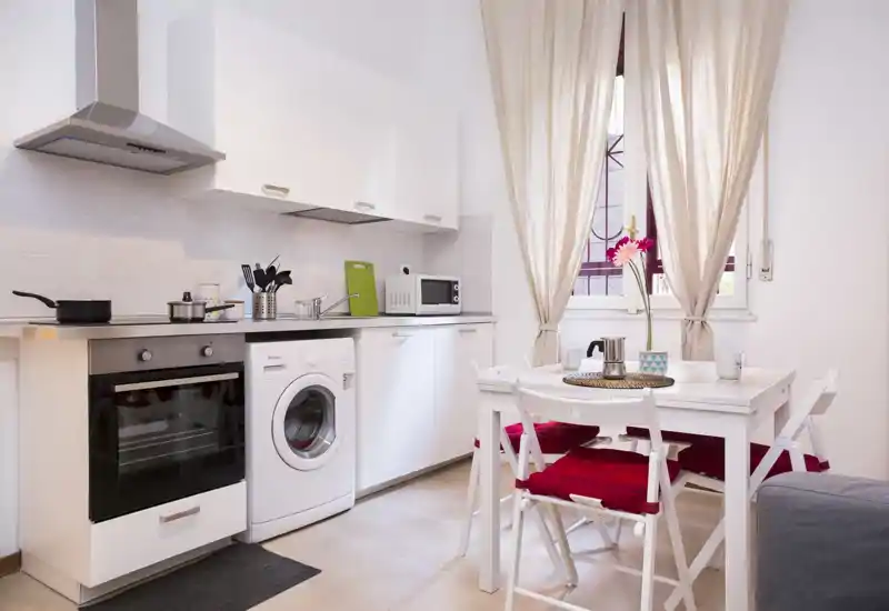 Compact modern kitchen with white cabinetry, built-in oven and washing machine, a small dining table with red cushions and natural light from a window — ideal for a cozy apartment.