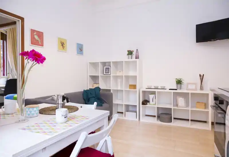 Bright, tidy living area with a dining table in the foreground, a cozy grey sofa, shelving units and a wall-mounted TV — ideal for a compact apartment.