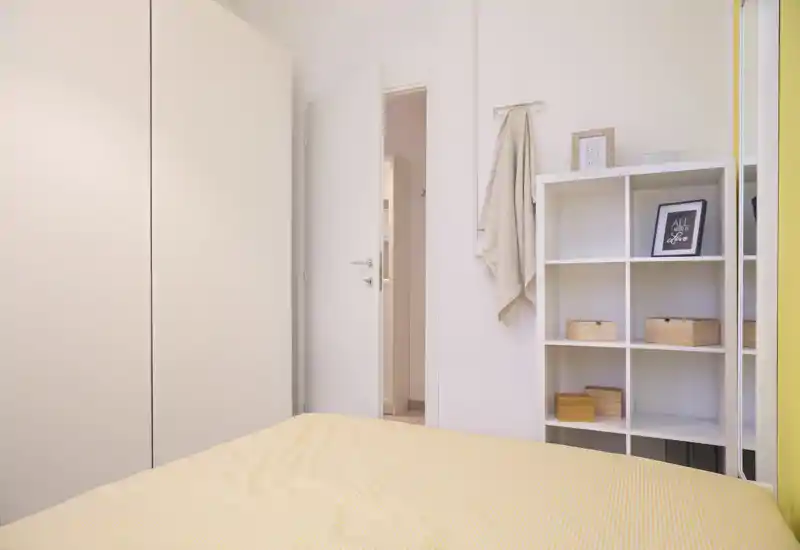Dormitorio luminoso y minimalista con funda amarilla, armario blanco y estantería de cubos; estilo moderno y luz suave que crean un espacio tranquilo.
