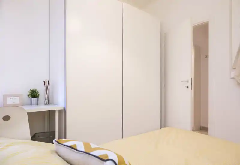 Dormitorio luminoso y acogedor con cama bien hecha de ropa de cama amarilla, un gran armario blanco y un pequeño escritorio junto a la ventana.