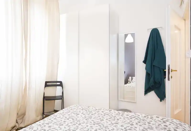 Camera da letto luminosa e compatta con una coperta a fantasia, armadio bianco alto e luce naturale morbida da una grande finestra. Stile semplice e ordinato, ideale per annunci.