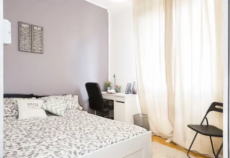 Camera da letto luminosa e ordinata con biancheria stampata, una scrivania e una grande finestra che diffonde luce naturale — accogliente e ben curata per annunci.