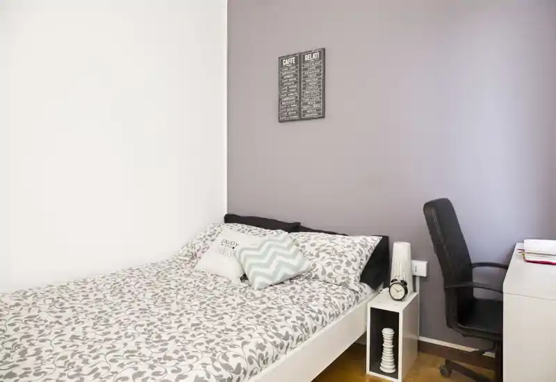 Camera da letto singola luminosa e ordinata con biancheria decorata, cuscini, comodino e una scrivania con sedia — ideale per uno studente o una persona sola.