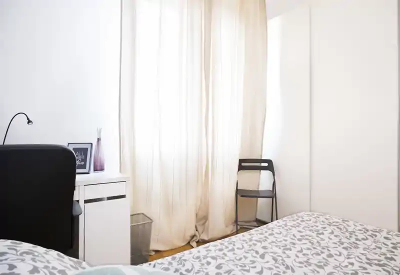 Rincón de dormitorio luminoso y de estilo suave con ropa de cama estampada, escritorio y silla junto a una gran ventana con cortinas translúcidas — ordenado y acogedor.