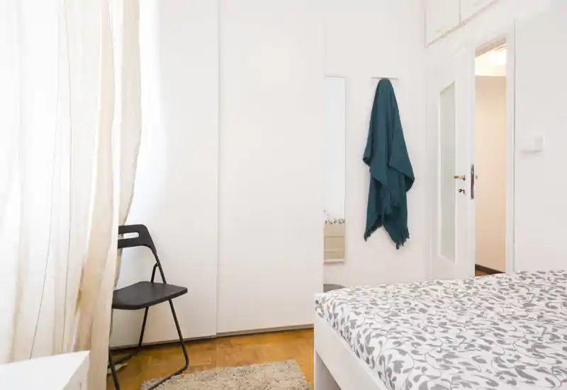 Camera da letto luminosa e minimal con piumone a fantasia, armadio e una sedia semplice vicino alla finestra — accogliente e ben illuminata per annunci.
