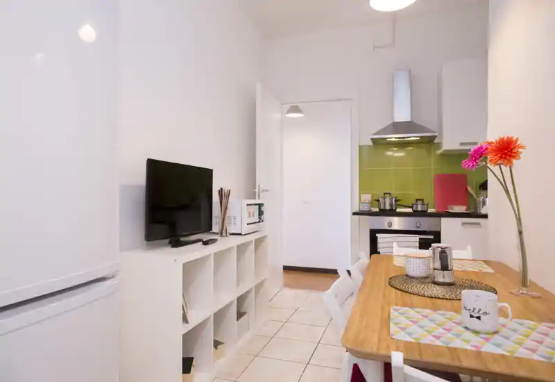Cocina pequeña y luminosa con una mesa de comedor. El espacio cuenta con placa y campana, frigorífico, microondas y una mesa de madera con manteles y flores, ideal para un piso en la ciudad.
