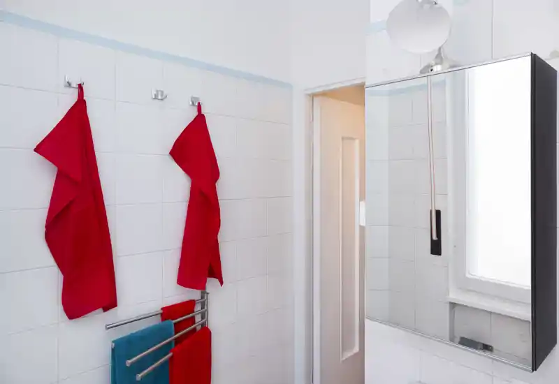 Baño luminoso y limpio con paredes alicatadas blancas, un armario con espejo y toallas de colores colgadas en ganchos y en el toallero.