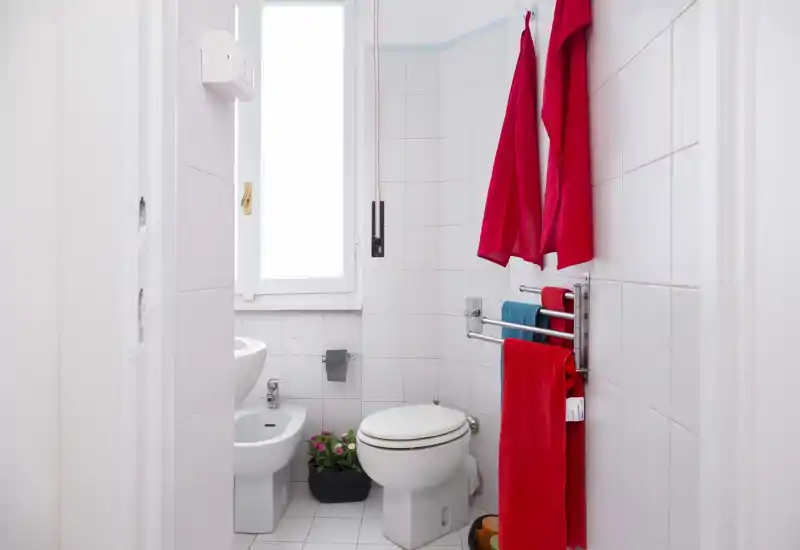 Baño compacto y luminoso con inodoro, bidé y lavabo; azulejos blancos y toallas de colores que dan un aspecto fresco y ordenado.
