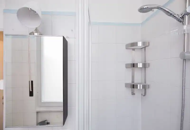 Baño alicatado y limpio con ducha de pared y estantes de esquina en acero inoxidable; se ve un armario con espejo y una pequeña ventana, aportando un área de ducha luminosa y funcional.