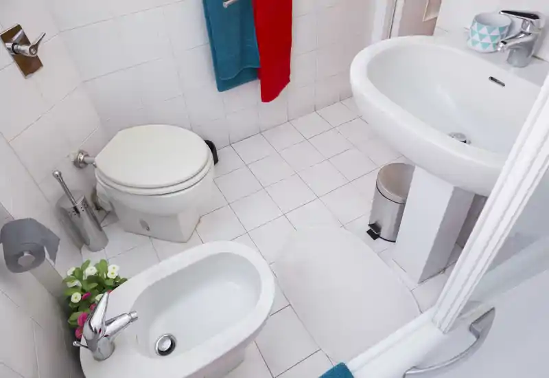 Baño compacto y limpio con azulejos blancos, lavabo de pedestal, inodoro y bidé; buena iluminación y orden.