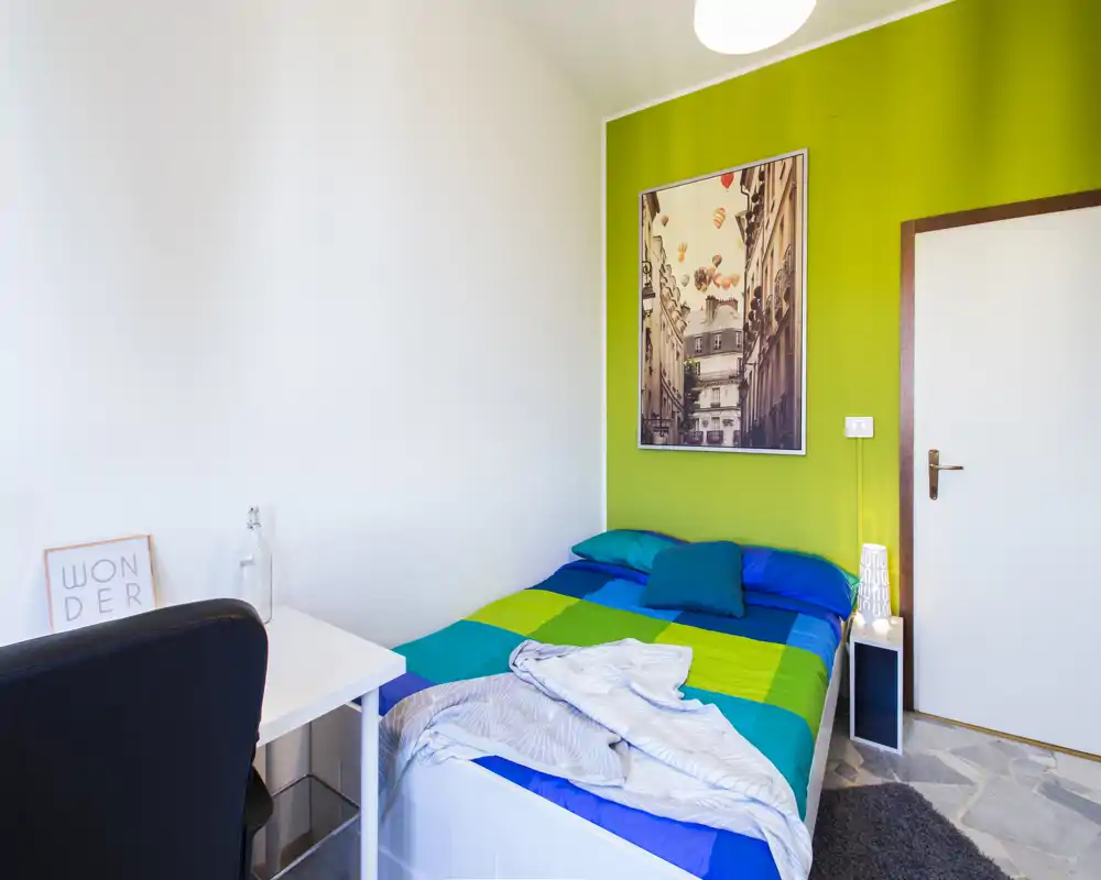 Bright, compact bedroom with a colorful double bed against a green accent wall, bedside lamp, small desk and simple wall art — ideal for a cozy single or couple stay.