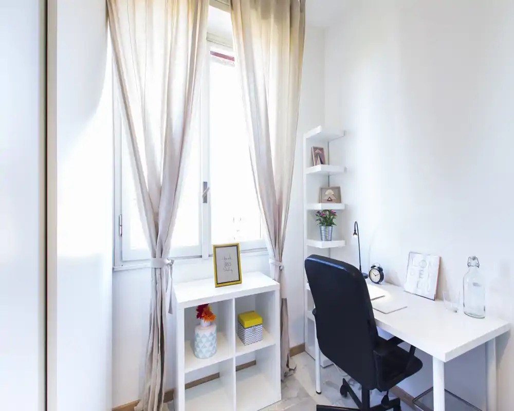 Bright, compact home-office corner with a white desk, ergonomic chair, shelves and soft curtains letting in natural light — ideal for remote work or study.