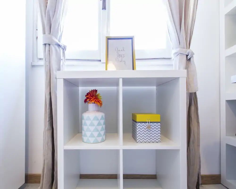 Image shows a close-up of a white open shelving unit with decorative items (vase with flowers, small box) positioned in front of a bright window and tied-back curtains. The photo focuses on decor and storage rather than a distinct room function.