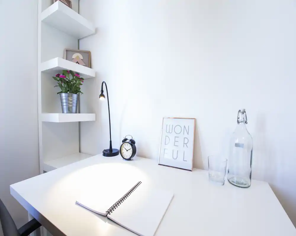 Bright, minimal desk area in a living/work corner with a lamp, notebook, small plant and decorative items — a clean, modern workspace within a living area.