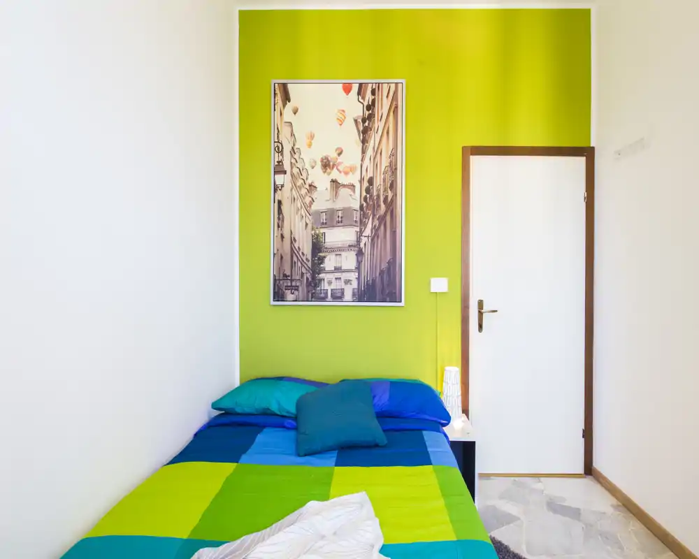 Bright, compact bedroom with a colorful double bed against a lime-green accent wall, decorative pillows and a framed print above the headboard.