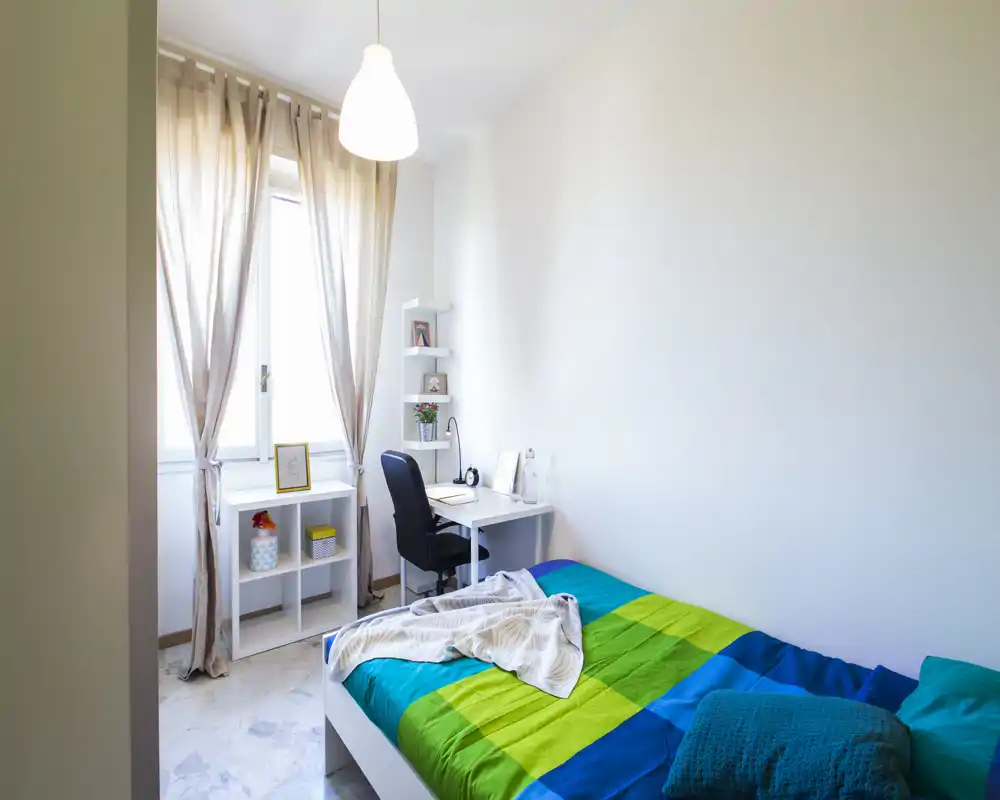 Bright, simply furnished single bedroom with a neatly made bed, desk and natural light from a large window. The room features light walls, a pendant lamp and small shelving next to the desk.