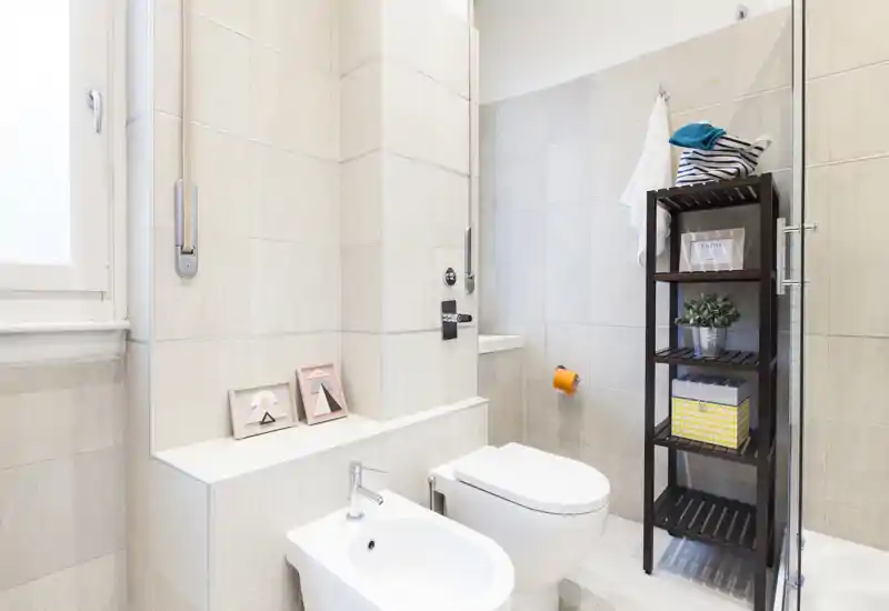 Bagno moderno e luminoso con WC, bidet e una sottile scaffalatura accanto alla doccia in vetro. Piastrelle dai toni neutri e luce naturale creano un ambiente pulito e arioso.